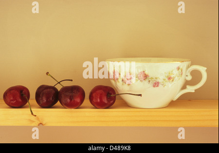 Augenhöhe Blick auf schmalen Holzregal mit vier dunklen roten Kirschen mit Stielen aufgereiht neben Phantasie weiß Teetasse Stockfoto