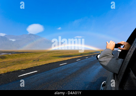 Mann in einem Auto schießt Süden isländische Landschaft unter einem strahlend blauen Himmel mit doppelter Regenbogen Stockfoto