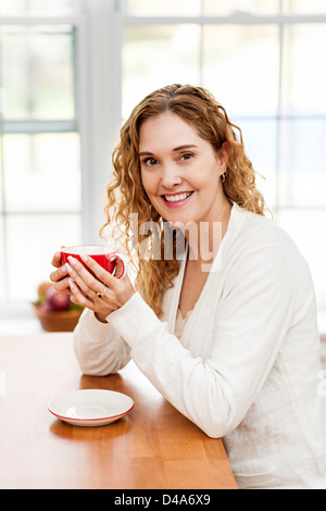 Porträt von lächelnden Frau halten rote Tasse Kaffee am Tisch in der Küche zu Hause Fenster Stockfoto