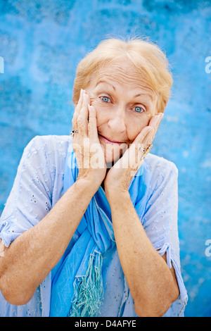 Alte blonde Frau Porträt, Dame in eleganter Kleidung mit Händen auf Gesicht vor Freude und Überraschung auf blauem Hintergrund Stockfoto