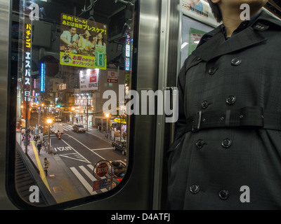 Mann im Trenchcoat, ein Passagier auf einer Yamanote Linie S-Bahn erhöht über einem langen Nacht Zeit Straße, Bahnhof Shin-Okubo, Tokio, Japan. Stockfoto