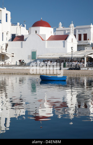 Weiße Kirche mit roter Kuppel, Cafés im Freien, Boot und Reflexionen im Wasser des Hafens von Mykonos, griechische Inseln Stockfoto