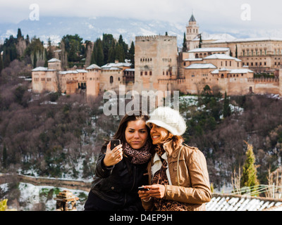 Zwei weibliche Touristen Freunde nehmen eine Selfie mit einer Handy-Kamera und schneebedeckten Alhambra-Palast im Hintergrund. Stockfoto