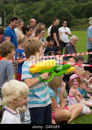 Masse der Menschen-Erwachsene und Kinder mit kleinen Jungen schießen große Kunststoff-Wasserpistole mit Menschen im Hintergrund Lachen UK Stockfoto