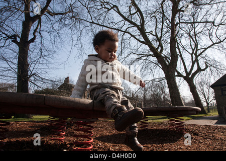 Kleinkind Jungen tragen Creme Jumper und Kampf Hose sitzt am Tisch im Park mit Bäumen hinter Stockfoto