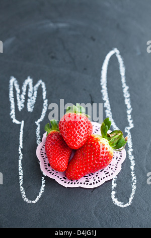 Drei ganze Erdbeeren und Kreidezeichnungen von Messer und Gabel auf dunklem Hintergrund Stockfoto