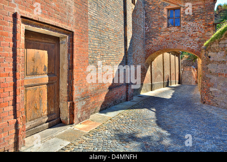 Schmalen gepflasterten Straße entlang roten Backsteinmauer mit alten Holztür und gewölbten Durchgang in Stadt von Guarene in Piemont, Italien. Stockfoto