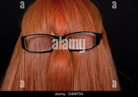 Mädchen mit langen roten Haaren bedeckte ihr Gesicht und das Tragen einer Brille vor einem schwarzen Hintergrund. Stockfoto
