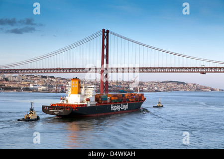 LISSABON, PORTUGAL. EIN CONTAINERSCHIFF MIT ZWEI SCHLEPPERN, LEITET DIE SALAZAR-BRÜCKE. AUCH GENANNT DER 25. APRIL-BRÜCKE UND DEN TEJO Stockfoto