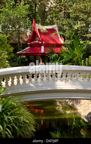 Thailand, Bangkok. Das Prasart Museum. Serenity Garden, Brücke über Gartenteich mit roten Thai Gebäude. Stockfoto