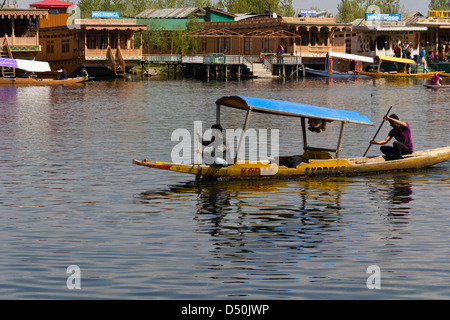 2 Männer paddeln Shikara in den ruhigen Gewässern des Sees Dal in Srinagar, mit Hausbooten im Hintergrund Stockfoto