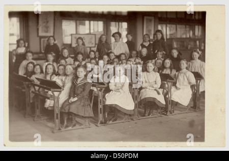 Junge Edwardian Mädchen mit verschränkten Armen, am Schreibtisch in einem Klassenzimmer sitzen, die Lehrer in den Hintergrund, in der Grundschule, ca. 1908, U.K. Edwardians Stockfoto