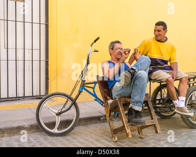 Zwei kubanische Männer sitzen vor einem hellen gelben Wand durch ihre Fahrräder gestikulieren und einem beheizten Gespräch in Havanna, Kuba Stockfoto