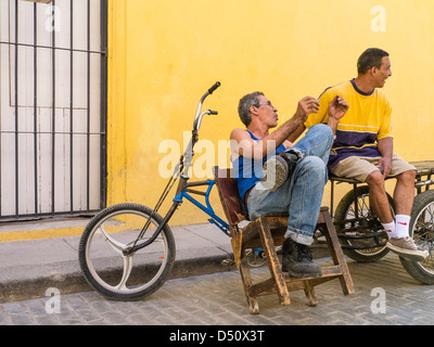 Zwei kubanische Männer sitzen vor einem hellen gelben Wand durch ihre Fahrräder gestikulieren und einem beheizten Gespräch in Havanna, Kuba Stockfoto