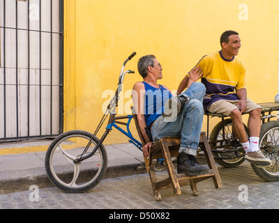 Zwei kubanische Männer sitzen vor einem hellen gelben Wand durch ihre Fahrräder gestikulieren und einem beheizten Gespräch in Havanna, Kuba Stockfoto