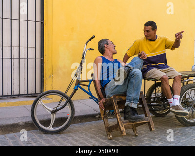 Zwei kubanische Männer sitzen vor einem hellen gelben Wand durch ihre Fahrräder gestikulieren und einem beheizten Gespräch in Havanna, Kuba Stockfoto