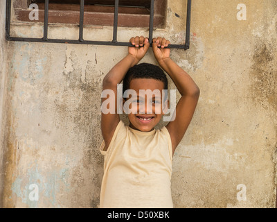 Eine 8-10 Jahre alten Hispanic kubanischen jungen hält an eine Eisen Fenster reiben über einem Fenster seines Hauses mit seinen Armen über dem Kopf. Stockfoto