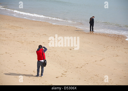 Menschen nehmen Foto von Mann mit langen Haaren gebunden Pferdeschwanz auf Handy ansehen Meer am Strand von Bournemouth im März Stockfoto