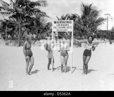 Junge Frauen, die sich lustig machen über Schild am Strand erfordert volle Badeanzüge: Miami, Florida Stockfoto