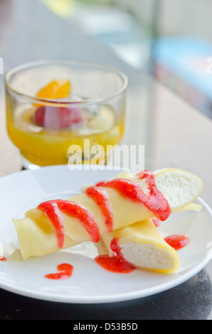 die Roll Pfannkuchen mit Sirup Erdbeeren und Sahne auf Teller und Mix Obst im Glas, Dessert auf Tisch Stockfoto