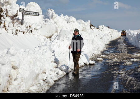 Janet Lees, ein Wanderer, steht an der Kreuzung der Cronk-y-Voddy und Peel Coast Road auf der Isle of man, während ein schwerer Bagger einen Weg durch tiefen Schnee im Hintergrund räumt. Stockfoto