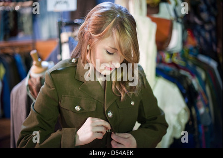 Hübsche junge Frau Tasten up und versucht auf eine alte grüne Armee-Jacke in einem Vintage-Kleidung fair mit Zahnstangen Kleidung hinter ihr Stockfoto