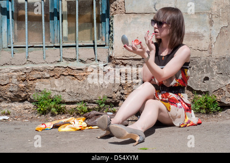 Das junge Mädchen sitzt auf Asphalt in der Nähe von der schmutzigen Haus aus Beton in den Sommernachmittag und macht Lippen Glanz Stockfoto