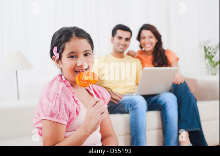 Mädchen essen einen Lolli mit ihren Eltern mit Laptop im Hintergrund Stockfoto