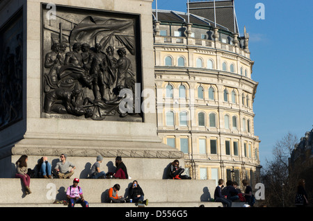 Die Frühlingssonne geniessen Touristen sitzen auf den Stufen rund um den Sockel der Nelsonsäule in Trafalgar Square. Stockfoto