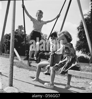 Historische, 1950. England, Sommer und drei kleine Kinder zusammen draußen auf einem traditionellen Metall- und Holzbrett oder 'Rock-A-Bye' Schwingen. Stockfoto