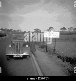 Historische, 1950er Jahre. Treiber stehen am Rande der Straße neben seinem Auto neben einem Schild "Willkommen in Schottland". Stockfoto