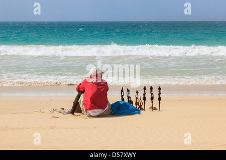 Lokalen junger schwarzer Mann sitzt in der heißen Sonne anzeigen afrikanischen Schnitzereien als Souvenirs am Strand von Boa Vista, Kap Verde Inseln zu verkaufen Stockfoto