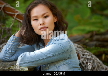 Schöne junge asiatische Frau tragen Jeans Hemd sitzt neben rustikalen Felswand vor der üppigen grünen Laub. Stockfoto