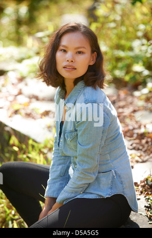 Schöne junge asiatische Frau tragen Jeans Hemd sitzt neben rustikalen Felswand vor der üppigen grünen Laub. Stockfoto
