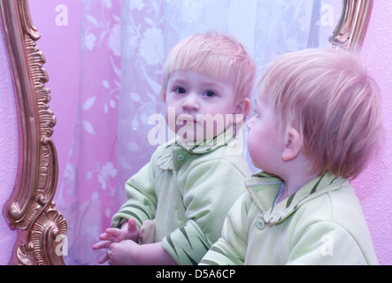 Das schöne Kind sucht eigene Reflexion im Spiegel, an der hell rosa Wand hängen Stockfoto