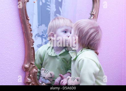 Das schöne Baby ist eine eigene Reflexion im Spiegel küssen, an der hell rosa Wand hängen Stockfoto