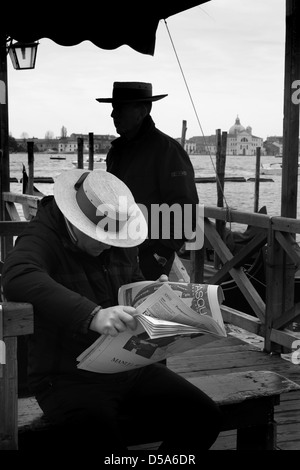 Schwarzweiß-Foto von zwei Gondolieri warten für touristische Kunden am Markusplatz, Venedig. Ein gondoliere im Hintergrund ab, der andere sitzt und liest eine Zeitung Stockfoto