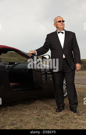 Ältere Mann mit schwarzen Anzug im Freien in Dünenlandschaft stehen auf der Rückseite seiner Sportwagen. Stockfoto