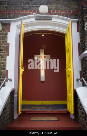 Ein Jesus rettet Neon-Schild am Eingang einer evangelischen Kirche in Peckham, Südlondon. Die gelbe Türen an der Spitze der Treppe mit zwei Fußmatten sind offen für Gläubige dieser christlichen Gemeinde in Südlondon begrüßen zu dürfen. Im Inneren befinden sich die Stimmen und die Schreie der Gläubigen, versammelten sich am Ostersonntag, ein wichtiges Datum im christlichen Kalender. Das Kreuz ist auf der Innenseite montiert Wand, durch die Neonröhre in der Kunststoff-Außenmantel beleuchtet. Stockfoto