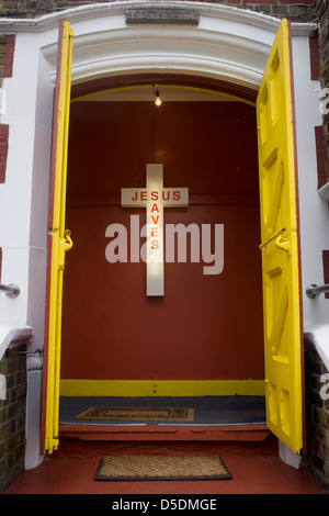 Ein Jesus rettet Neon-Schild am Eingang einer evangelischen Kirche in Peckham, Südlondon. Die gelbe Türen an der Spitze der Treppe mit zwei Fußmatten sind offen für Gläubige dieser christlichen Gemeinde in Südlondon begrüßen zu dürfen. Im Inneren befinden sich die Stimmen und die Schreie der Gläubigen, versammelten sich am Ostersonntag, ein wichtiges Datum im christlichen Kalender. Das Kreuz ist auf der Innenseite montiert Wand, durch die Neonröhre in der Kunststoff-Außenmantel beleuchtet. Stockfoto