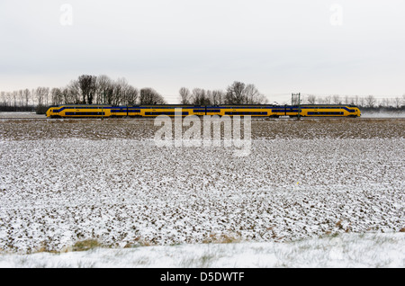 Vorbei an gelben Zug der niederländischen Eisenbahnen im Schnee bedeckt Landschaft. Zeeland, Niederlande. Stockfoto