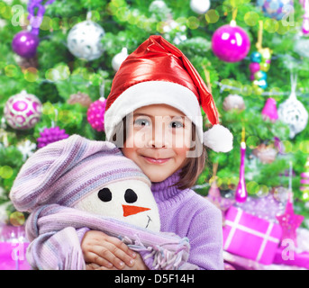 Porträt von kleinen hübschen Mädchen tragen rote Weihnachtsmütze umarmt Stofftier weiße Schneemann über Weihnachtsbaum, Silvester-party Stockfoto