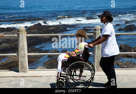 Senior-Frau im Rollstuhl wird geschoben entlang Sea Point Promenade durch Betreuer - Kapstadt - Südafrika Stockfoto