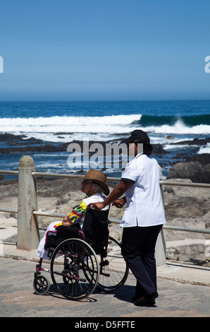 Senior-Frau im Rollstuhl wird geschoben entlang Sea Point Promenade durch Betreuer - Kapstadt - Südafrika Stockfoto