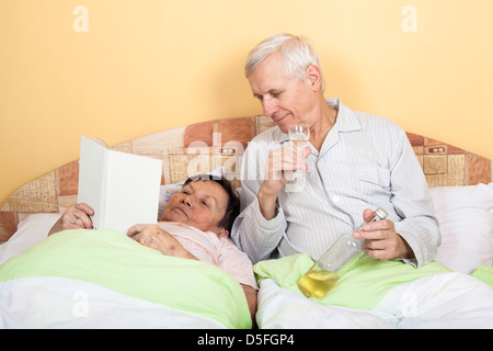 Lustiger senior Mann trinken Alkohol und Frau liest Buch im Bett Stockfoto