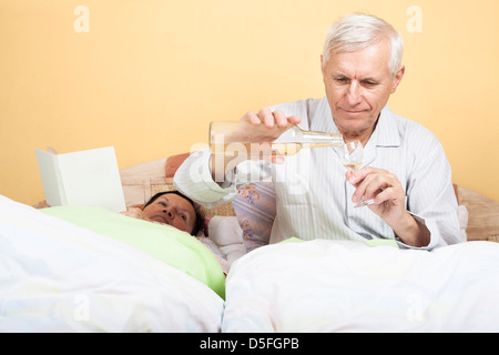 Senior mit Flasche Alkohol und Frau Mann ältere Menschen mit Buch im Bett Stockfoto