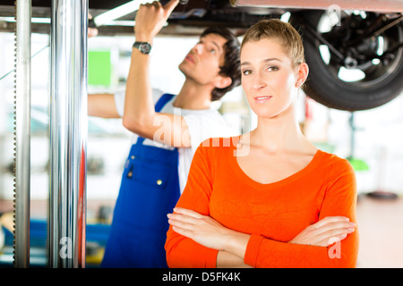 Frau stand vor ihrem Auto, das auf einem Auto Hebebühne steht, macht eine Reparatur unter dem Auto Mechaniker aufgehoben wird Stockfoto