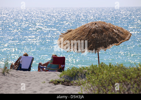 Eine braune Tahiti Stil Rasen Sonnenschirm und zwei Personen sitzen auf Stühlen an einem Strand am Meer Stockfoto