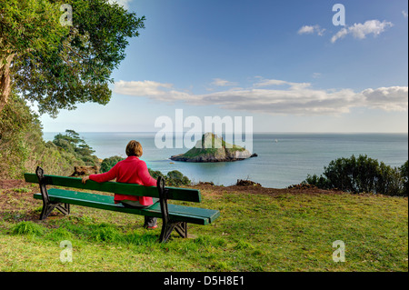 Frau in Rot entspannt auf einer Bank mit Blick auf die Ansicht von Thatcher rock, einem beliebten Aussichtspunkt, in Torquay, Devon, Großbritannien Stockfoto