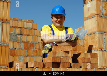 Mason tragen gelbes T-shirt und blaue Helm mit Edelstahl-Kelle posiert unter unfertige Haus Ziegelwände Stockfoto